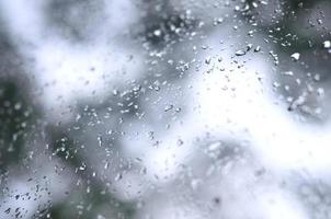 ein Foto von Regentropfen auf dem Fensterglas mit verschwommenem Blick auf die blühenden grünen Bäume. abstraktes Bild, das bewölkte und regnerische Wetterbedingungen zeigt