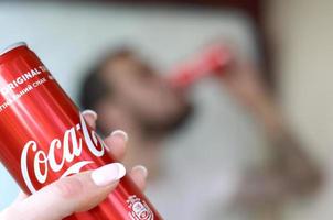 Charkow. ukraine - 2. mai 2019 kaukasischer mann trinkt coca-cola-getränk im garageninnenraum und männliche hand präsentiert eine rote coca-cola-dose im fokus foto