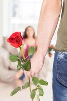 Überraschung für sie. Zugeschnittenes Bild eines Mannes, der eine rote Rose hält, während seine Freundin im Hintergrund sitzt foto
