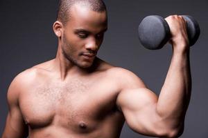 Gewichtheben. junger hemdloser afrikanischer mann, der mit hantel trainiert, während er vor grauem hintergrund steht foto