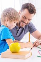 Elternunterstützung. Seitenansicht des fröhlichen jungen Vaters, der seinem Sohn hilft, Hausaufgaben zu machen, während er zusammen mit grünem Apfel im Vordergrund am Tisch sitzt foto