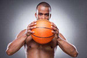 Basketball ist mein Leben. Junger hemdloser afrikanischer Mann, der einen Teil seines Gesichts hinter einem Basketballball versteckt und in die Kamera schaut, während er vor grauem Hintergrund steht foto