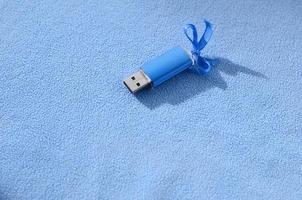 Eine leuchtend blaue USB-Flash-Speicherkarte mit einer blauen Schleife liegt auf einer Decke aus weichem und pelzigem hellblauem Fleece-Stoff. klassisches weibliches Geschenkdesign für eine Speicherkarte foto