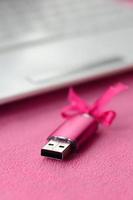 Eine leuchtend rosa USB-Flash-Speicherkarte mit einer rosa Schleife liegt auf einer Decke aus weichem und pelzigem hellrosa Fleece-Stoff neben einem weißen Laptop. klassisches weibliches Geschenkdesign für eine Speicherkarte foto