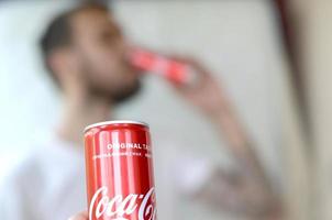 Charkow. ukraine - 2. mai 2019 kaukasischer mann trinkt coca-cola-getränk im garageninnenraum und männliche hand präsentiert eine rote coca-cola-dose im fokus foto