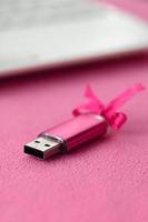 Eine leuchtend rosa USB-Flash-Speicherkarte mit einer rosa Schleife liegt auf einer Decke aus weichem und pelzigem hellrosa Fleece-Stoff neben einem weißen Laptop. klassisches weibliches Geschenkdesign für eine Speicherkarte foto