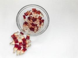 flaschenförmige Gummibärchen liegen in einem durchsichtigen, runden Teller. andere gummis werden in der nähe gegossen. auf einem weißen matten Hintergrund. appetitlich und süß, leckeres Dessert foto