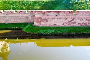 Eine große Erdmauer aus rotem Backstein ist stark schützend und ein Wassergraben mit dem Krieg einer alten, alten mittelalterlichen Burg in der Mitte Europas gegen den blauen Himmel foto