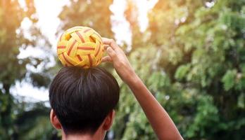 Junger südostasiatischer männlicher Sepak-Takraw-Spieler, der mit seiner rechten Hand den Ball auf dem Kopf hält, Sepak-Takraw-Spiel im Freien nach der Schule, weicher und selektiver Fokus auf den Ball.
