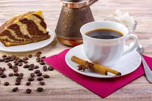 eine tasse schwarzen kaffee auf untertasse mit leckerem kuchen. weiße zuckerwürfel in einer schüssel, teelöffel, anis, zimtstangen und anderem zubehör für den kaffee auf dunklem holzhintergrund. foto