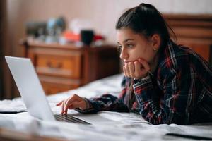 junge Frau arbeitet, während sie mit Laptop auf dem Bett liegt foto