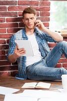 beschäftigt zu Hause arbeiten. Schöner junger Mann, der auf dem Handy spricht und auf sein digitales Tablet schaut, während er auf dem Hartholzboden in seiner Wohnung sitzt foto