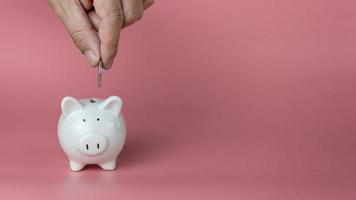 Eine menschliche Hand lässt eine Münze in ein weißes Sparschwein fallen. auf einem rosa Hintergrund. Spar- und Anlagekonzepte. foto