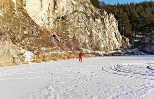 Junger Mann in roter Winterkleidung, der entlang des zugefrorenen schneebedeckten Flusses in der Nähe schöner Klippen Ski fährt. winteraktivitäten, sport, aktiver lebensstil foto