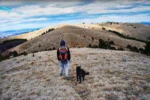Junger Mann von hinten in einer Kapuze und mit einem Rucksack, der mit einem schwarzen Hund vor einem blauen bewölkten Himmel in den Hügeln spazieren geht foto