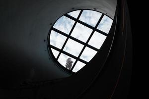Fenster im Dunkeln, rundes Glasfenster im Korridor, Boden im Tunnel, U-Bahn-Innenarchitektur, Mann steht auf der runden Glasdecke vor blauem Himmel und weißen Wolken, transparente Kuppel. foto