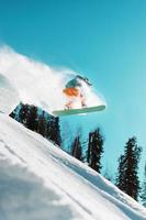 Snowboarder springt von einem hohen schneebedeckten Sprungbrett in einem Wald vor blauem Himmel foto