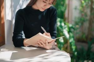 asiatische berufstätige frau, die schwarzes hemd trägt und tagebuch auf kleinem notizbuch auf dem tisch im innencafé schreibt. Frau stellt fest und trinkt Kaffee im Café. Arbeiten von überall Konzept. foto