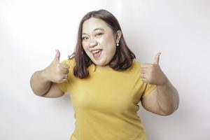 Aufgeregte große asiatische Frau mit gelbem Hemd gibt Daumen nach oben Handgeste der Zustimmung, isoliert durch weißen Hintergrund foto