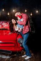 junge schöne eltern, die ihre kleine süße tochter in den armen halten und spaß in der nähe von retro-autos im studio haben. weihnachtlicher Familienlook. landschaft des neuen jahres. foto