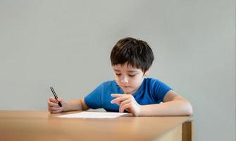 Kind, das auf dem Tisch sitzt und Hausaufgaben macht, Kind, Junge, der schwarzen Stift hält und auf weißem Papier schreibt, kleiner Junge, der zu Hause englische Wörter übt. Grundschule und Hausunterricht, Fernunterrichtskonzept foto