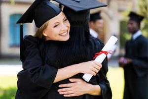 Herzlichen Glückwunsch zwei glückliche Frauen in Graduierungskleidern, die sich umarmen und lächeln, während zwei Männer im Hintergrund stehen foto