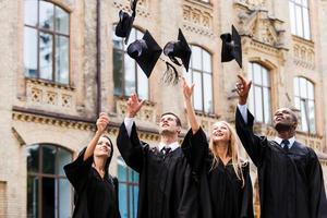 Wir haben endlich unseren Abschluss gemacht, vier glückliche College-Absolventen in Abschlusskleidern, die ihre Doktorwürste werfen und lächeln, während sie in der Nähe der Universität stehen foto