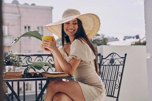 Fröhliche junge Frau mit elegantem Hut, die ein heißes Getränk genießt, während sie sich auf dem Balkon entspannt