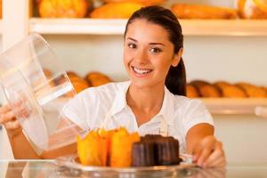 Probieren Sie diese attraktive junge Frau in Schürze, die einen Teller mit frischen Keksen trägt und lächelt, während sie in der Bäckerei steht foto