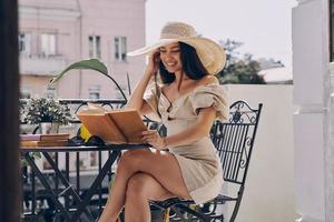 attraktive junge Frau in elegantem Hut liest Buch und lächelt beim Entspannen auf dem Balkon foto