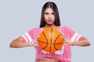Bist du bereit, eine selbstbewusste junge Cheerleaderin zu spielen, die einen Basketballball hält und in die Kamera schaut, während sie vor grauem Hintergrund steht? foto