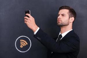 Wo ist ein Wi-Fi-Bild eines hübschen jungen Mannes, der ein Telefon hält und nach einer Internetverbindung sucht, während er gegen das Teilen von Symbolkreidezeichnung auf der Tafel steht foto
