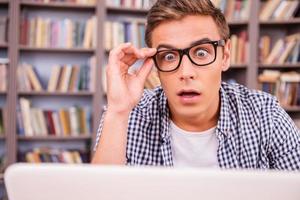Es ist unglaublich überrascht, wie ein junger Mann auf einen Laptop schaut und den Mund offen hält, während er gegen ein Bücherregal sitzt