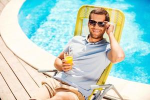 dies ist die lebensansicht eines fröhlichen jungen mannes im poloshirt, der am liegestuhl sitzt und ein glas mit orangensaft hält foto