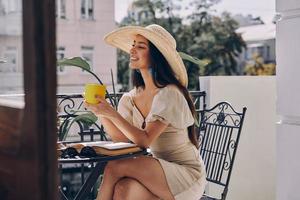 Attraktive junge Frau mit elegantem Hut, die ein heißes Getränk genießt und lächelt, während sie sich auf dem Balkon entspannt