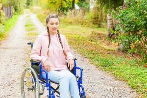 junge glückliche Frau mit Handicap im Rollstuhl auf der Straße im Krankenhauspark, die auf Patientendienste wartet. gelähmtes Mädchen im Rollstuhl für Behinderte im Freien in der Natur. Rehabilitationskonzept. foto