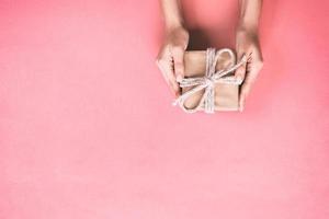 Frauenhände geben eingewickeltes Valentinsgruß- oder anderes Feiertagsgeschenk in Papier mit rosa Schleife. geschenkbox, dekoration des geschenks auf rosa tisch, draufsicht mit kopierraum foto