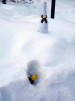 Eine weiße Flasche Champagner, die im weißen, frostigen Schnee vergraben war, und ein gelber Flaschenverschluss, einer wurde in der Nähe begraben. sei bereit für partyfeiern, weihnachtsgrüße, frohe feiertage, weihnachten, neujahr foto