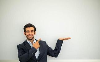 porträt eines lächelnden jungen kaukasischen geschäftsmannes, der einen dunklen anzug trägt und kopienraum auf seinen handflächen präsentiert und auf die betonwand zeigt. foto