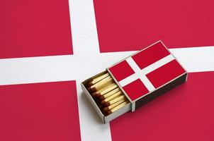 Die dänische Flagge wird in einer offenen Streichholzschachtel gezeigt, die mit Streichhölzern gefüllt ist und auf einer großen Flagge liegt foto