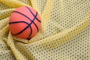 kleiner orangefarbener gummibasketball liegt auf einer gelben sportjersey-kleidungsstoffstruktur und einem hintergrund mit vielen falten foto