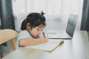 asiatisches baby, das ein blau gestreiftes hemd trägt, benutzt laptop und schreibt notizen in notizbuch, um online auf holztischschreibtisch im wohnzimmer zu hause zu lernen. bildung lernen online von zu hause aus.