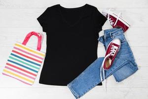 T-Shirt schwarz und Turnschuhe. t-shirt-modell flach lag mit sommeraccessoires. Jeans und Turnschuhe auf Holzbodenhintergrund. Platz kopieren. Vorlage leere Leinwand. Vorderansicht von oben. foto