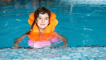 Ein Kind in einer orangefarbenen Schwimmweste schwimmt im Pool in einem Aquakomplex für Familienferien. Sicherheit auf dem Wasser, Schwimmen lernen foto