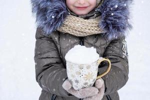 Ein Mädchen in warmer Winterkleidung hält einen Becher mit Schnee in den Händen - kalte Wetterbedingungen, Saisonalität. weihnachten und neujahr foto