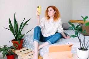 Junges rothaariges Mädchen macht ein Selfie zu Hause auf dem Sofa auf einem Smartphone foto