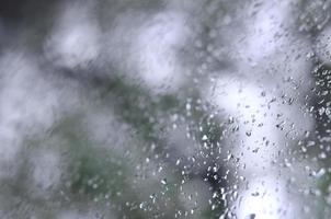 ein Foto von Regentropfen auf dem Fensterglas mit verschwommenem Blick auf die blühenden grünen Bäume. abstraktes Bild, das bewölkte und regnerische Wetterbedingungen zeigt