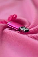 Die leuchtend rosa USB-Flash-Speicherkarte mit rosa Schleife liegt auf einer Decke aus weichem und pelzigem hellrosa Fleecestoff mit vielen Relieffalten. Speichergerät im Damendesign foto