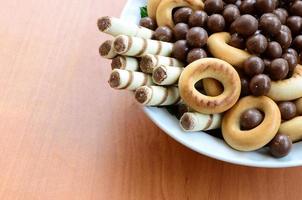 knusprige röhrchen, schokoladenkugeln und bagels liegen in einem weißen teller auf einem holztisch. Mischung aus verschiedenen Süßigkeiten foto