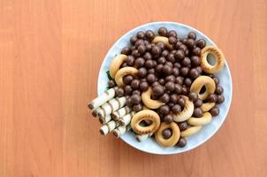 knusprige röhrchen, schokoladenkugeln und bagels liegen in einem weißen teller auf einem holztisch. Mischung aus verschiedenen Süßigkeiten foto